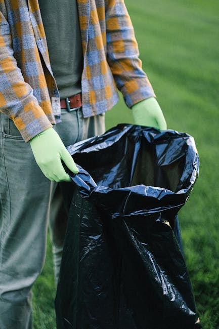A person wearing a checkered shirt with shades of yellow, blue, and gray, along with lime green gloves, is holding open a large black plastic rubbish bag outdoors on a grassy area. The individual is standing on a well-maintained lawn, and part of their waist and arms are visible, with the background showing lush green grass. The black rubbish bag is empty, open at the top, and appears ready for waste collection. The scene suggests an outdoor waste disposal activity, possibly related to garden or household waste removal. The person's attire and the use of gloves imply a focus on hygienic rubbish handling. This visual aligns with services such as private rubbish collection offered by companies like Rubbish Collection Ilford, providing alternative waste handling solutions outside of local authority disposal options.
