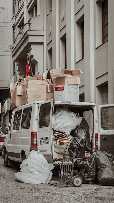 Photograph of a white commercial van parked on a city street next to a multi-storey building with a light grey façade. The van's rear doors are open, revealing a large collection of waste materials inside, including black plastic rubbish bags, flattened cardboard boxes, and assorted packaging debris. Several cardboard boxes are stacked on the roof of the van, some partially collapsed or torn, with visible corrugated surfaces and brown kraft paper finishes. The surrounding environment features a narrow sidewalk with additional rubbish bags placed at the rear of the vehicle and an empty hand trolly leaning against the side, indicating on-site waste collection or private refuse disposal. The background includes the building's windows and architectural details such as windowsills, ledges, and façade textures, suggesting an urban setting. Natural light illuminates the scene, highlighting the materials' textures and colours, aligned with waste management and rubbish removal services like those provided by Rubbish Collection Ilford, who specialise in alternative waste handling options.