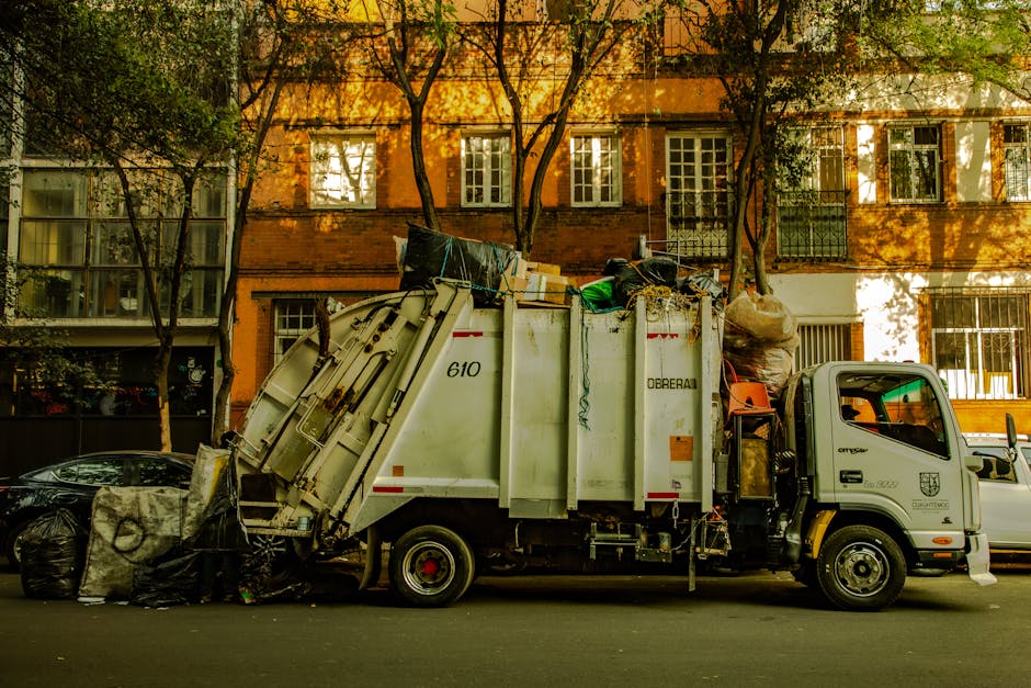 A street scene in daylight features a white waste collection truck loaded with a variety of collected rubbish, including black plastic garbage bags, cardboard boxes, plastic containers, and miscellaneous debris. The truck, parked parallel to a sidewalk, has visible red and orange reflective strips along its side and the number '610' printed on its cargo section, indicating a private rubbish collection service such as Rubbish Collection Ilford. The surrounding environment includes a row of multi-story residential buildings with a brick and plaster facade, multiple windows with white frames, and small balconies protected by metal railings. Tall, leafy trees aline the street, casting shadows across the buildings and vehicle, contributing to an atmospheric, daytime urban setting. The street surface is asphalt, and there are other parked vehicles, including a black car partially visible at the front left and a white van behind the truck. The scene emphasizes on-site waste collection, an alternative to traditional bin collections, with equipment and materials typical for commercial or private rubbish removal services.
