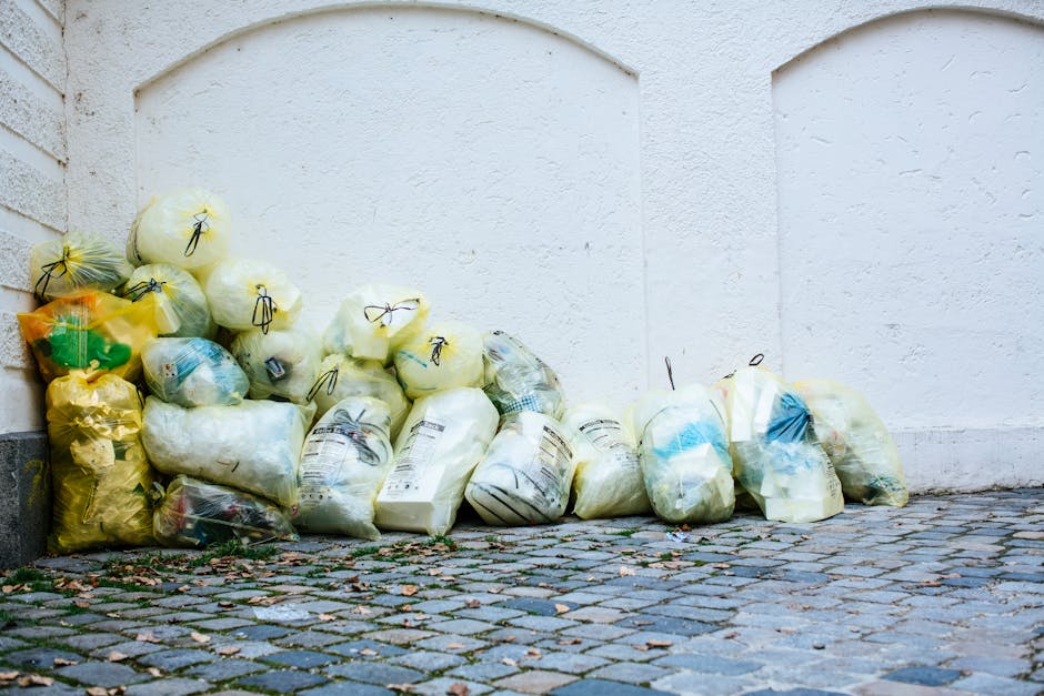 Photograph of a white commercial van parked on a city street next to a multi-storey building with a light grey façade. The van's rear doors are open, revealing a large collection of waste materials inside, including black plastic rubbish bags, flattened cardboard boxes, and assorted packaging debris. Several cardboard boxes are stacked on the roof of the van, some partially collapsed or torn, with visible corrugated surfaces and brown kraft paper finishes. The surrounding environment features a narrow sidewalk with additional rubbish bags placed at the rear of the vehicle and an empty hand trolly leaning against the side, indicating on-site waste collection or private refuse disposal. The background includes the building's windows and architectural details such as windowsills, ledges, and façade textures, suggesting an urban setting. Natural light illuminates the scene, highlighting the materials' textures and colours, aligned with waste management and rubbish removal services like those provided by Rubbish Collection Ilford, who specialise in alternative waste handling options.