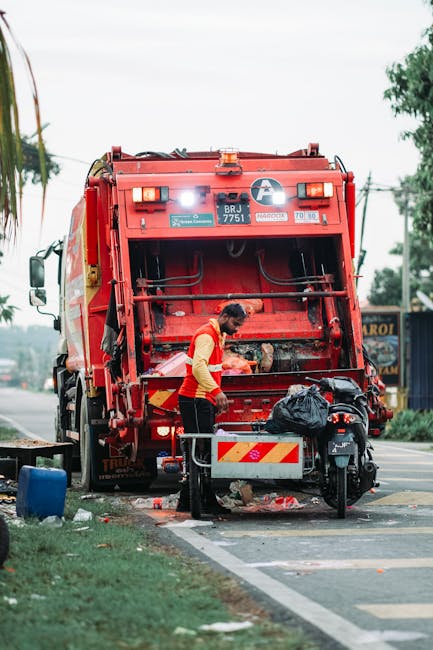 A waste collection worker wearing a high-visibility orange vest and dark trousers is standing beside a large red rubbish collection truck on a roadside. The worker appears to be operating controls or inspecting the back of the truck, which is designed for collecting and compacting waste. The truck is positioned on a paved road with a white and red reflective safety panel at the rear. To the side of the worker, there is a small black motorcycle parked close to the truck, with a black garbage bag placed on the motorcycle's seat or cargo area. The scene is outdoors, with greenery and trees in the background, along with some utility poles and wires, indicating an urban or suburban environment. The sky is overcast, and the overall atmosphere suggests a routine rubbish collection operation, possibly part of a private waste management or on-site clearance service, reflecting alternative waste handling options separate from local authority collection. The company's branding is not visible on the truck, but the context aligns with professional waste removal services such as those provided by Rubbish Collection Ilford.