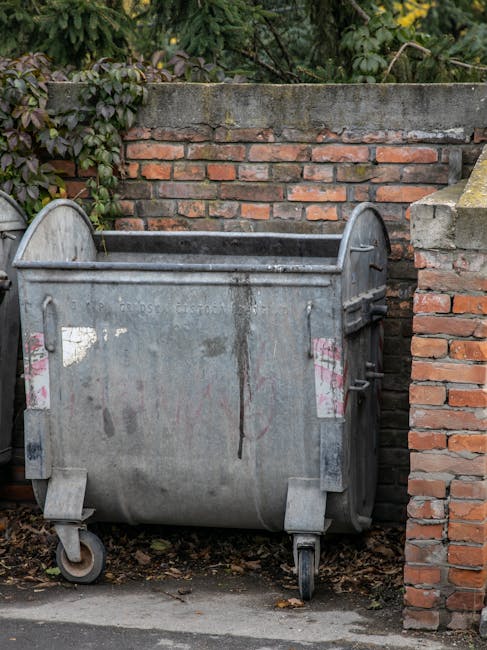 A large public metal waste skip positioned on a paved surface, situated between a brick wall and a concrete boundary wall. The skip is constructed from weathered, grey galvanized steel with visible patches of dirt, rust, and peeling paint, indicating frequent outdoor use. It features two sturdy wheels at the base for mobility, with side handles for manoeuvrability. Behind the skip, dense green foliage and leafy plants can be seen above the boundary wall, suggesting a garden or residential setting. The top edge of the skip is open, revealing an empty interior intended for collection and disposal of bulky waste or rubbish. The surrounding environment includes natural daylight illuminating the scene, and the overall setting appears to be part of an external space managed by organisations such as Rubbish Collection Ilford, providing an example of private waste handling options outside council collection services.