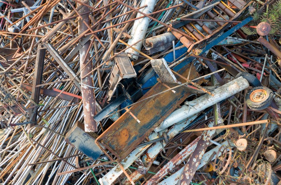 A large pile of discarded metal debris consisting of rusted steel pipes, bent and overlapping, with some tubular sections and welded joints visible. The materials display various shades of brown, orange, and grey corrosion, with some pipes having flaking paint or rough, oxidized surfaces. Among the pile, there are flattened metal sheets, some with bent edges and peeling paint, alongside a scattered selection of smaller metal fragments and hardware. The debris appears to be outdoors, situated on a paved or concrete surface, with a background of greenery partially visible through gaps in the metal. The lighting is natural, suggesting daylight, which highlights the textures and weathered appearance of the scrap materials. This scene reflects the type of waste collected during independent metal or scrap removal services provided by companies such as Rubbish Collection Ilford, emphasizing the importance of proper disposal or recycling of old, scrap metal objects.