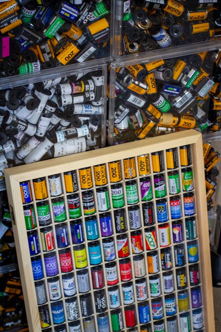 The image displays a collection of small paint and marker containers organized in a wooden display case with multiple compartments, each filled with various coloured caps indicating different shades of paint or ink. Behind the display, there are clear plastic boxes filled with numerous similar cylindrical containers, predominantly black, white, yellow, and green, with labels and colour coding visible on the caps. The containers appear to be made of plastic, with some caps showing a matte finish and others a slight sheen, reflecting ambient lighting. The setting appears to be a storage or supply area, possibly within a workshop or art supply store, with an emphasis on organisation for easy selection. The scene is well-lit, highlighting the vibrant colours and details of the labels, which include numerical values and branding. This image relates to proper organisation and storage of art or craft supplies, connecting to waste management or disposal services that could handle surplus or unwanted containers as part of an alternative waste handling process, provided by Rubbish Collection Ilford.