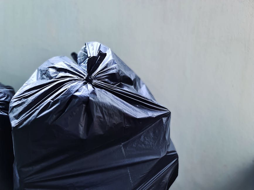 Close-up image of black plastic rubbish bags filled with waste, pulled tightly and secured with knots at the top, resting against a plain light-colored wall. The bags appear to be thick, glossy, and durable, with some creases and reflections visible on their surface, indicating they are made of standard refuse sack material. The primary focus is on the stacked or gathered bags, positioned on a flat surface such as a driveway or pavement, with minimal background detail. The lighting is neutral, showing the bags’ textures and the slightly crinkled appearance of the plastic. The scene likely depicts an interim step in waste collection or disposal, such as an on-site rubbish clearance, consistent with private waste handling services offered by Rubbish Collection Ilford. The image emphasizes the typical appearance of ready-for-collection waste bags in a professional rubbish removal context.