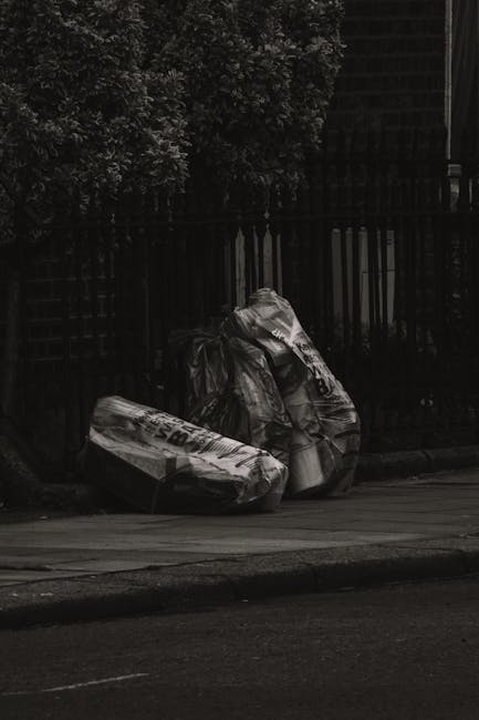 The image shows two large black plastic garbage bags filled with waste, placed on the edge of a paved sidewalk next to a curb. The bags appear to be tightly packed and sealed, with some creases and slight tears visible on the surface. They are positioned in front of a dark metal fence with vertical bars, which runs parallel to the sidewalk. Behind the fence, dense foliage of leafy trees is visible, with some branches extending above the fence line. The scene is lit with natural light, casting subtle shadows on the pavement and the bags. The setting appears to be an outdoor urban area, possibly on a residential street, where refuse has been collected for disposal. The context suggests this could be part of an independent rubbish collection service, similar to those offered by companies like Rubbish Collection Ilford, operating in the area. The overall atmosphere is quiet and undisturbed, with no other visible activity or vehicles present.