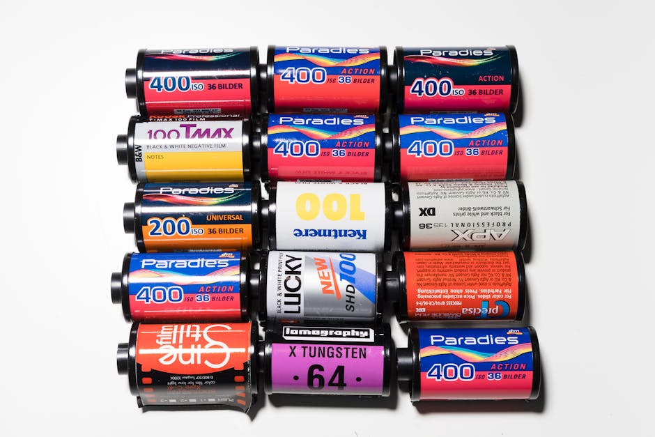 A collection of various rolls of photographic film arranged on a white surface, with some positioned horizontally and others vertically. The rolls are primarily black, white, and red, with labels displaying different brand names, film speeds, and specifications such as '400 ISO' and '100', along with other technical details. The textures of the film rolls are smooth with glossy labels that reflect light slightly. The background is plain white, emphasizing the cylindrical shapes and vibrant label colors. This array of film rolls is displayed in a neat, organized manner, possibly for storage or display purposes, and subtly relates to the concept of preservation or archiving, which can be associated with document or material disposal services such as those offered by Rubbish Collection Ilford in the context of alternative waste handling and on-site clearance for sensitive items.
