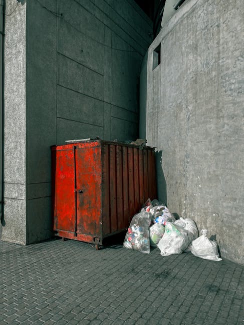 A red metal dumpster with visible rust and signs of wear, positioned against the corner of a building with dark green paneling on one side and a light grey concrete wall on the other. Several white plastic bags filled with waste are placed around the base of the dumpster, some leaning against it. The bags appear to contain mixed household rubbish, with plastic bottles and other small debris visible through the semi-transparent material. The ground is paved with dark grey interlocking bricks, and natural daylight illuminates the scene, highlighting the textures of the rusted metal and the rough concrete surfaces. The setting suggests a background of private or commercial waste disposal, possibly related to on-site rubbish clearance managed by a waste removal service such as Rubbish Collection Ilford, offering an alternative to council rubbish collection.