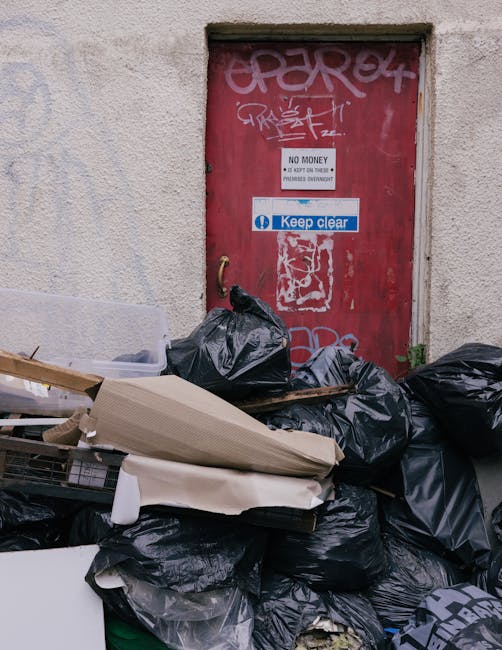 A large pile of black rubbish bags, mixed with cardboard and other refuse, is stacked against a rough-textured, off-white exterior wall of a building. The bags are crumpled and tightly packed, with some partially torn open, revealing waste inside. The pile extends to the foreground and overlaps a small piece of white and brown packaging material. Behind the bags, there is a red metal door with visible graffiti in pink and white. The door features a white sign stating 'No Money' and an additional smaller notice beneath it, along with a blue sticker that reads 'Keep clear.' The wall surrounding the door appears weathered, with slight discoloration and patches of dirt. The scene suggests an outdoor area prone to unmanaged waste accumulation, possibly associated with private rubbish collection services like those offered by Rubbish Collection Ilford, illustrating the need for professional rubbish removal or on-site clearance to manage excess waste effectively.