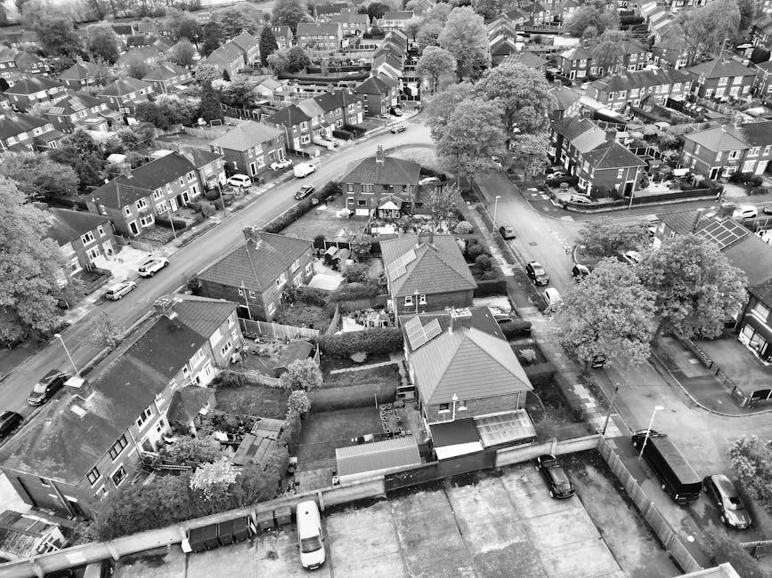 An aerial view of a residential area showing multiple detached and semi-detached houses with pitched roofs, some with tiled finishes and others with metal or slate coverings. The houses are arranged along curved streets with parked cars lining the roads and driveways, including a white van and several smaller vehicles. In the foreground, there is a backyard with a neatly maintained garden, separated by a low hedge, featuring a small shed, patio area, and garden furniture. Surrounding trees provide partial shade, and a few properties have solar panels installed on roofs. The environment appears organized with clear delineation between individual properties, and the overall scene captures a typical suburban neighbourhood in daytime, with light and shadow suggesting overcast weather. This image relates to private waste management or rubbish collection, highlighting the kind of area serviced by waste removal specialists like Rubbish Collection Ilford, who support residents with rubbish disposal and alternative waste handling solutions outside local authority arrangements.