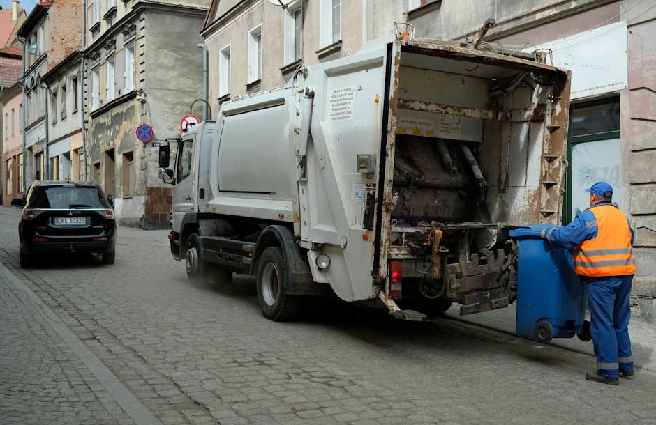 A row of waste collection bins and a large metal storage container situated outdoors on a paved surface, with dense foliage visible in the background. The waste bins are plastic, with some labeled for specific waste types such as paper and general rubbish, and are positioned side-by-side, with lids closed. The metal container is positioned behind the bins, featuring sliding doors, a weathered finish, and warning or service labels attached to its surface. The scene is illuminated by natural light, casting subtle shadows on the ground and on the containers. The overall setting suggests an area designated for private waste disposal or alternative rubbish removal, managed by a waste management company like Rubbish Collection Ilford, rather than a municipal recycling point. The environment reflects a typical outdoor refuse collection site, with functional storage for different types of waste awaiting collection or disposal, aligning with the service context of rubbish removal solutions in Ilford.