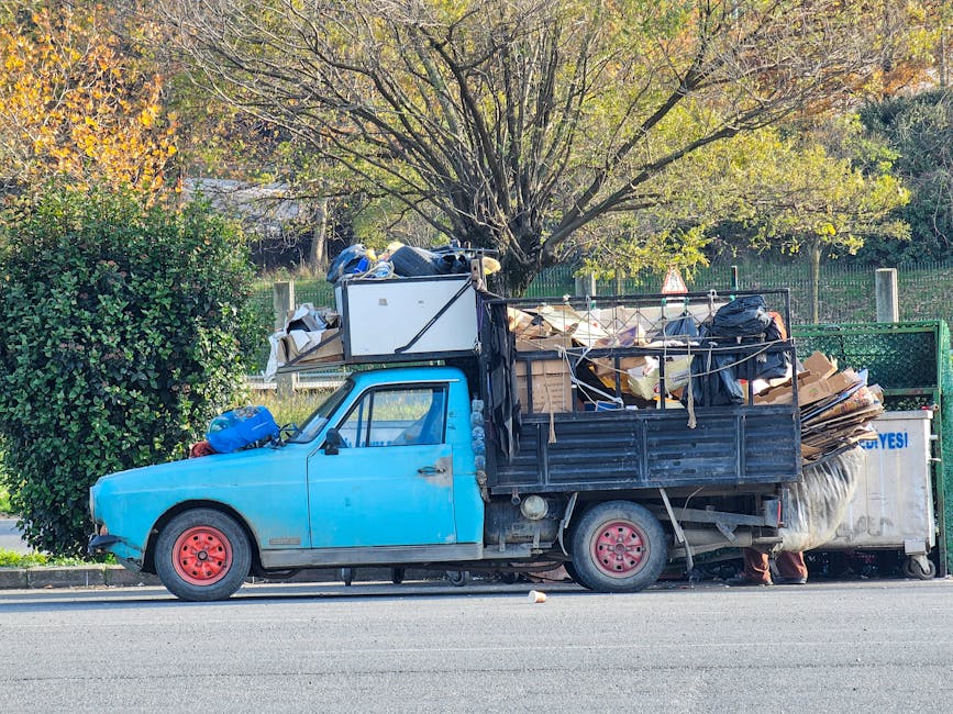 A small, blue, vintage pickup truck parked on the side of a street, loaded with a large, overflowing collection of mixed waste materials, including cardboard boxes, black plastic bags, and various debris, secured with ropes attached to the truck’s black metal side panels. The truck has visible red wheels, some of which are worn and faded, and a passenger side mirror. In the background, there is a large tree with green and yellow leaves, indicating an autumn setting, alongside a green metal fence, with a grassy area beyond. The truck appears to be used for private waste collection or on-site clearance and is positioned on a paved road with minimal traffic. The scene is well-lit with natural daylight, reflecting an environment where independent rubbish removal services, such as those provided by Rubbish Collection Ilford, might operate in the context of alternative waste handling outside of local authority rubbish collection. The image captures the practicality of a waste vehicle used for rubbish removal in an urban or suburban setting, emphasizing the volume of waste typical of bulky waste or furniture disposal services.