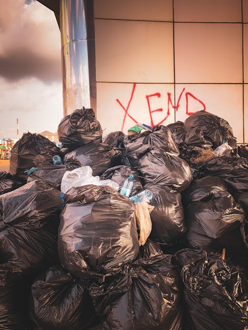 A large pile of tightly tied black plastic rubbish bags is situated outdoors against a tiled wall. The bags vary in size and are stacked haphazardly, with some leaning against the wall and others piled on top. Littered among the bags are clear plastic water bottles, some partially crushed and others appearing to be full. The surface beneath the bags is not visible, obscured by the accumulation. In the background, the wall features graffiti with the word 'XEND' spray-painted in red. To the left, part of a metallic vertical structure reflects light, and a cloudy sky with hints of sunlight is visible above, suggesting late afternoon or early evening. This scene exemplifies a stubborn accumulation of waste that could relate to non-standard or private waste collection services, such as those offered by companies like Rubbish Collection Ilford, especially during delays or disruptions in municipal rubbish collection schedules.